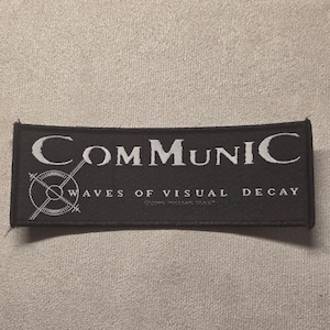 May include: A rectangular black embroidered patch featuring the white text COMMUNIC and WAVES OF VISUAL DECAY. To the left of the text is a white circular graphic with crosshairs and radiating lines. The patch has a stitched border.