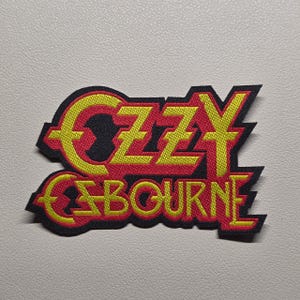 Może przedstawiać: Haftowana naszywka z napisem OZZY OSBOURNE stylizowaną czcionką. Litery są jaskrawożółte z wyraźnym czerwonym obrysem, umieszczone na czarnym tle, które podąża za konturem tekstu, tworząc ostry i wyraźnie zdefiniowany kształt naszywki.