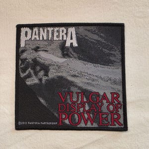 May include: A square fabric patch featuring the Pantera logo in white at the top and the text Vulgar Display of Power in red lettering at the bottom. The background displays a grayscale graphic design with a copyright notice in the corner.