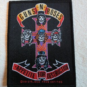 May include: A rectangular black embroidered patch featuring the Guns N' Roses logo with a central cross design. The cross displays five skull figures, with a banner above reading Guns N' Roses and a banner below reading Appetite For Destruction.