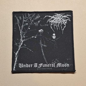 May include: A square black fabric patch featuring a white graphic print of a tree and a figure. The text at the top reads Darkthrone in a stylized font, while the bottom text reads Under A Funeral Moon and 2010 DARKTHRONE in white lettering.