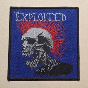 May include: A square embroidered patch with a blue background featuring a white skull with a red mohawk hairstyle. The text THE EXPLOITED is printed in white above the skull. The patch has a thick black border around the entire square edge.