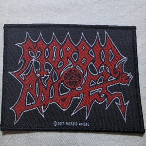 Op de afbeelding: Een rechthoekige zwarte stoffen patch met het rode Morbid Angel-logo in een gestileerd, grillig lettertype. Een cirkelvormig embleem met een pentagramontwerp is in de tekst gecentreerd. De tekst 2017 MORBID ANGEL staat onderaan in het wit.