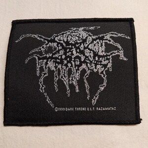 May include: A rectangular black fabric patch featuring a white embroidered graphic design with intricate, flowing lines. The text "©1999 DARK THRONE U.L.T. RAZAMATAZ" is printed in white at the bottom center of the patch, which has a stitched border.