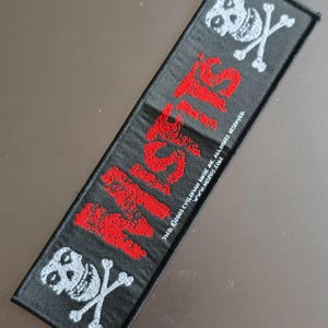May include: A rectangular black embroidered patch featuring the red text MISFITS oriented vertically. The patch includes a white skull and crossbones graphic at both the top and bottom ends, along with small white copyright text along the side.