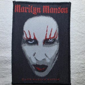 May include: A rectangular embroidered patch featuring a black background with the text Marilyn Manson in red at the top. The centre displays a stylised, pale face with red markings above the eyes and dark lips. The text 2015 Marilyn Manson is at the bottom.