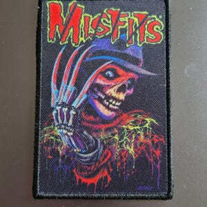 May include: A rectangular embroidered patch featuring the red text Misfits at the top. Below the text is a graphic of a skeletal figure wearing a hat with a clawed hand raised near its face. The design uses vibrant red, blue, and yellow colours.