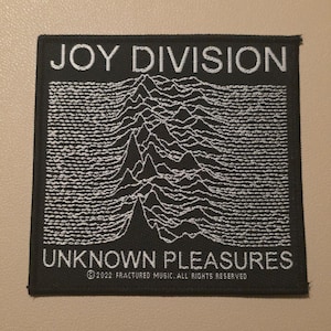 May include: A square black fabric patch featuring white embroidered text that reads JOY DIVISION at the top and UNKNOWN PLEASURES at the bottom. The centre displays a white line graph of radio waves. Text at the bottom reads 2022 FRACTURED MUSIC.