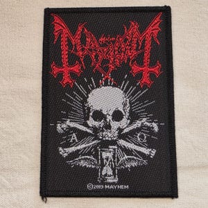May include: A rectangular black embroidered patch featuring the red Mayhem logo at the top. Below, a white skull sits above crossed bones, an hourglass, and wings. The Greek letters Alpha and Omega are on either side, with 2019 MAYHEM text at the base.