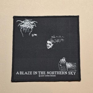 May include: A square black fabric patch featuring white embroidered graphics including a stylized tree, a face, and a hand. The text reads A BLAZE IN THE NORTHERN SKY and 2009 DARKTHRONE. The patch has a black stitched border and a woven texture.