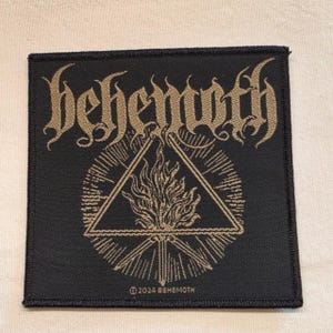 May include: A square black embroidered patch featuring the word behemoth in a stylised tan font at the top. Below the text is a tan graphic of a flame inside a triangle, surrounded by radiating lines. The text 2024 BEHEMOTH is at the bottom.