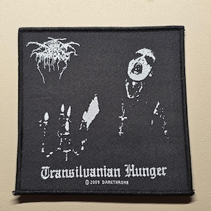 May include: A square black fabric patch featuring white embroidered graphics. The design includes a stylized logo, a candelabra, and a figure with an open mouth. The text reads Transilvanian Hunger and 2009 DARKTHRONE in white lettering at the bottom.
