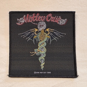 May include: A square black embroidered patch featuring the red text Mötley Crüe at the top. The centre design shows a silver skull atop a gold sword, wrapped in a green snake with black wings. The bottom text reads ©2018 MOTLEY CRUE.