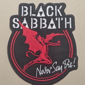 May include: A black embroidered patch featuring the white text BLACK SABBATH at the top with red winged figure in the center and the white script text Never Say Die! at the bottom, all enclosed within a circular red and black border.