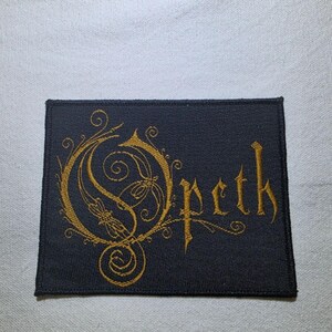 May include: A rectangular black embroidered patch featuring the word Opeth in a stylized gold font. The letter O is large and ornate with intricate swirling flourishes and decorative lines, while the remaining letters are in a gothic script.