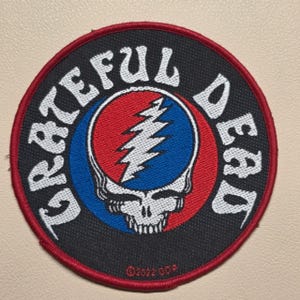 May include: A circular embroidered patch featuring a black background with a red border. The text GRATEFUL DEAD is stitched in white around the edge. In the center is a blue and red circular design with a white lightning bolt skull graphic.