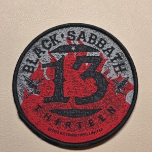 May include: A circular embroidered patch featuring the text Black Sabbath at the top and Thirteen at the bottom. The centre displays a large black number 13 over a red flame-like background with small winged figures on each side and text.