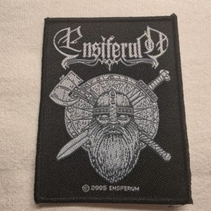 May include: A rectangular black embroidered patch featuring the white text Ensiferum at the top. The design depicts a bearded warrior wearing a helmet in front of a circular shield, with an axe and a sword crossed behind it. The text 2005 Ensiferum is at the bottom.