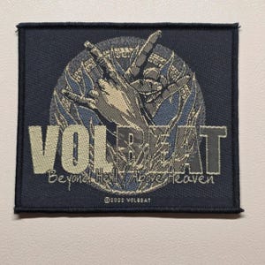 Peut inclure: Un écusson carré brodé noir représentant une main faisant un geste rock-and-roll sur un motif circulaire bleu et gris. Le texte indique VOLBEAT, Beyond Hell / Above Heaven et 2022 VOLBEAT en fil beige clair.