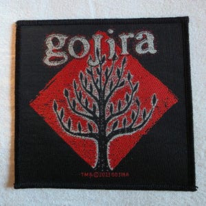 Può includere: Una toppa quadrata ricamata nera con una forma a rombo rossa al centro e la sagoma di un albero nero. La parola "gojira" è ricamata in grigio sopra il rombo e il testo "TM & © 2023 GOJIRA" è riportato in piccole lettere rosse.