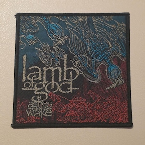 May include: A square fabric patch featuring a woven graphic design in shades of blue, red, and black. The text reads "lamb of god" and "ashes of the wake" in a stylised font. The design includes abstract figures and patterns with a black border.