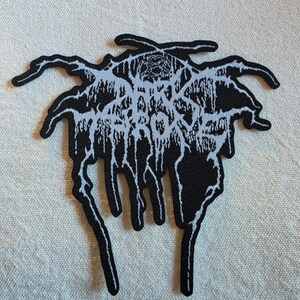 May include: A black and white embroidered patch featuring a stylized, jagged, and dripping font design that reads Darkthrone. The text is rendered in a sharp, intricate, and symmetrical style with long, thin, downward-reaching tendrils.
