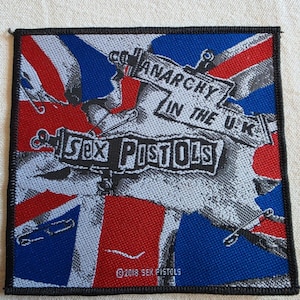 May include: A square embroidered patch featuring a red, white, and blue Union Jack flag background. The design includes the text ANARCHY IN THE U.K. and SEX PISTOLS in black lettering, accented with safety pin illustrations and a 2018 copyright.