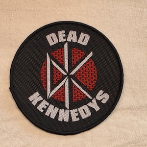 May include: A circular embroidered patch with a black background and a red circular center. The patch features the white text DEAD KENNEDYS arched around a central white logo consisting of four intersecting lines forming a stylized DK symbol.