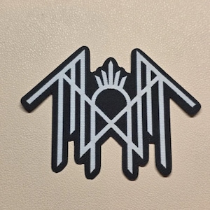 May include: A black embroidered patch featuring a white geometric design. The pattern consists of a central arch topped with a sunburst motif, flanked by two symmetrical wing-like structures with sharp, angular lines pointing outward.