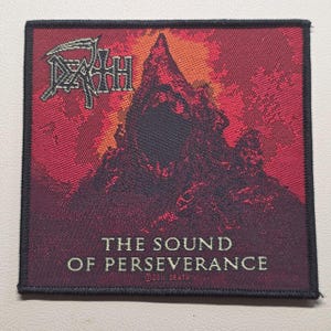 Op de afbeelding: Een vierkante stoffen patch met een donkerrode en zwarte bergafbeelding met een centrale leegte. Links bovenaan staat het woord DEATH in een gestileerd lettertype met een zeis, en de onderste tekst luidt THE SOUND OF PERSEVERANCE ©2011 DEATH.