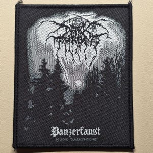 May include: A rectangular black and white embroidered patch featuring a forest landscape with a full moon and a stylized logo at the top. The text "Panzerfaust" is printed in white at the bottom, followed by the small text "© 2010 DARKTHRONE".