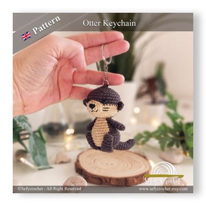 May include: A crocheted otter keychain with brown and beige fur. The otter is holding a silver key ring. The keychain is on a wooden surface.