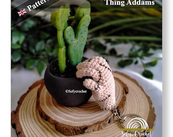Puede incluir: Una mano tejida a crochet con puntadas, de color rosa claro, con una línea negra cosida que recorre el centro de la mano. La mano está unida a un llavero plateado con un anillo redondo plateado. La mano descansa sobre una superficie de madera junto a una planta de cactus en maceta. El texto "Thing Addams" está escrito en letras negras sobre fondo blanco en la esquina superior derecha de la imagen. El texto "Sefy-Crochet" está escrito en letras negras sobre fondo blanco en la esquina inferior derecha de la imagen. El texto "www.sefycrochet.etsy.com" está escrito en letras negras sobre fondo blanco debajo del texto "Sefy-Crochet".