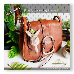 Puede incluir: Un bolso de cuero marrón con un charm de conejo tejido. El charm es beige con un trébol verde y está sujeto al asa del bolso. El texto "Lucky Bunny Bag Charm" es visible.