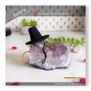 Può includere: Un portachiavi a forma di cappello coreano nero all'uncinetto con un dettaglio di perline, esposto su un cristallo viola. Il portachiavi è attaccato a un anello di legno. L'immagine include il testo "Pattern" e "Korean Hat Keychain".