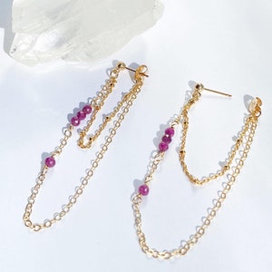 Peut inclure: Une paire de boucles d'oreilles en chaîne dorée avec de petites perles violettes. Les boucles d'oreilles ont deux chaînes qui pendent du lobe de l'oreille.
