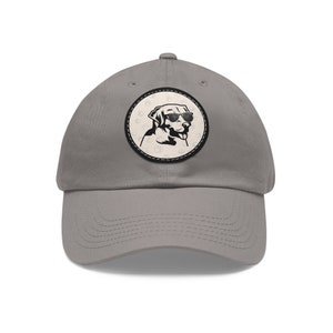 May include: A grey baseball cap with a circular patch. The patch features a black and white illustration of a dog wearing sunglasses, set against a beige background with paw prints. The cap has a curved peak and an adjustable strap.
