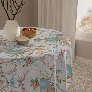 May include: A round white tablecloth with a floral pattern featuring pink roses, blue ribbons, and gold accents. The design includes ornate chair motifs. A white vase, a bowl, and a basket of apples are on the table.