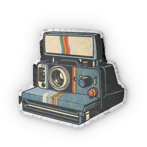 May include: A vintage-style pillow shaped like a Polaroid camera. The camera is primarily blue with rainbow stripes and a retro design. The pillow has a white border and a distressed, aged appearance.