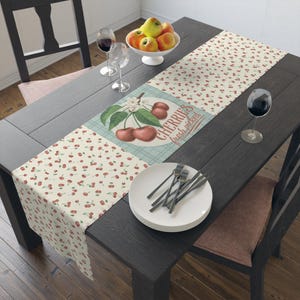 May include: A dark wood dining table set with a cherry-themed table runner, white plates, and silverware. A bowl of apples sits on the table. Wine glasses and a chair are also visible.