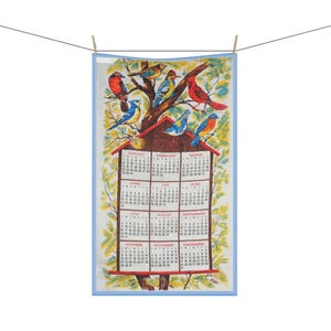 Könnte beinhalten: Ein dekoratives Kalenderhandtuch mit einer farbenfrohen Illustration verschiedener Vögel, die auf einem Baum mit einem Vogelhaus sitzen. Das Kalendarium ist sichtbar, mit Monaten und Daten. Das Handtuch ist mit einer hellblauen Bordüre versehen und hängt an einer Wäscheleine.