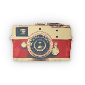 May include: A decorative pillow shaped like a vintage camera. The pillow features a cream-colored top and a red bottom, with a detailed lens and buttons. The design evokes a retro aesthetic.