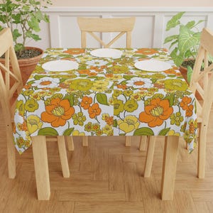 Puede incluir: Un mantel rectangular con un estampado floral retro en naranja, amarillo, verde y blanco. El mantel está sobre una mesa de madera, con tres platos blancos. El tema general es la decoración del hogar vintage.