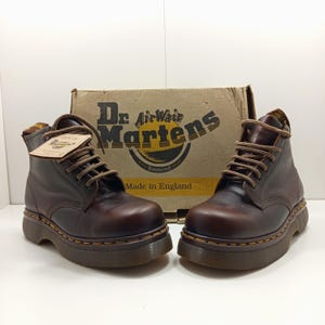 Può includere: Un paio di stivaletti Dr. Martens in pelle marrone con lacci marroni e cuciture gialle. Gli stivali sono esposti davanti a una scatola di cartone con il logo Dr. Martens e la scritta "Made in England".