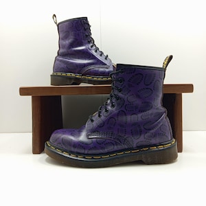 May include: A pair of purple leather boots with a black lace-up design. The boots feature a repeating pattern of black imprints and a yellow trim around the sole. One boot is on a small wooden shelf, and the other is below.