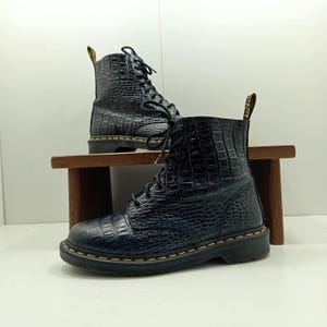 May include: Black leather boots with a crocodile skin texture. The boots have a lace-up design, yellow stitching, and a black sole. One boot is in the foreground, and another is in the background, both on a wooden surface.