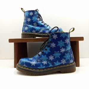 May include: A pair of blue leather boots with a snowflake pattern. The boots have black laces, yellow stitching, and a thick sole. The boots are displayed on a small wooden shelf.