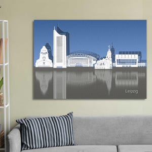 Könnte beinhalten: Ein Leinwanddruck der Skyline von Leipzig in Weiß vor blauem Himmel. Die Gebäude spiegeln sich im Wasser darunter. Das Wort "Leipzig" ist unten rechts in Weiß gedruckt.