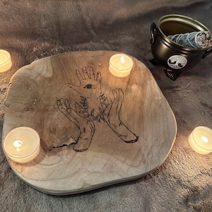 Könnte beinhalten: Ein Holztablett mit einem handgezeichneten Design aus zwei Händen mit einem Auge in der Mitte. Das Tablett ist von vier weißen Teelichtern umgeben.