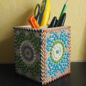 Op de afbeelding: Een mozaïek potloodhouder met een blauw, groen en wit patroon. De houder is gevuld met verschillende schrijfbenodigdheden, waaronder een schaar, een gele pen en een paar potloden.